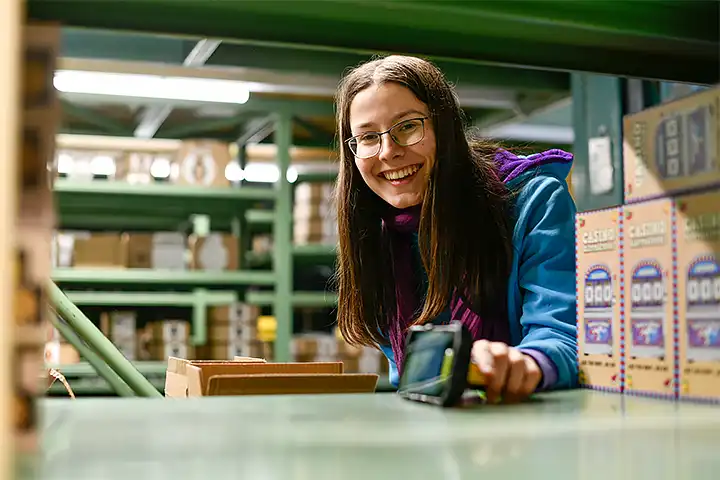 Junge Logistikerin lächelt durch ein Regal bei der Arbeit